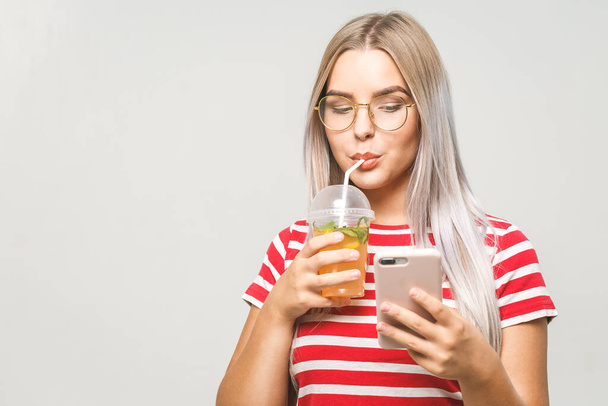 Beautiful young woman drinking a homemade detox juice, texting on her phone while isolated over white. Using phone.  - Photo, Image