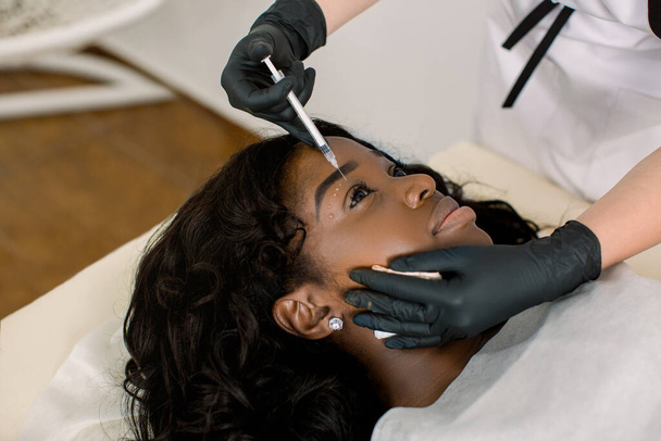 Close up of hands of doctor cosmetologist making the rejuvenating facial injections procedure for tightening and smoothing wrinkles on the face skin around the eyes for beautiful young African woman - Zdjęcie, obraz