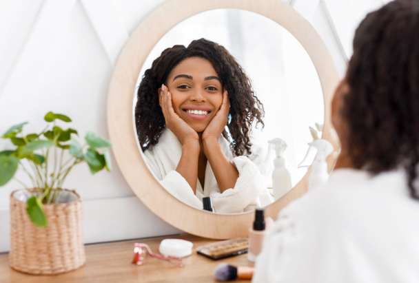 Hermosa mujer negra en bata de baño sentada en la mesa de vestir, mirando al espejo
 - Foto, imagen