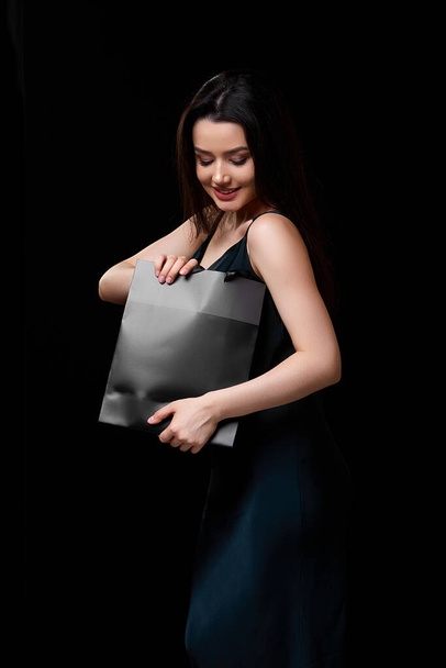 a young beautiful woman in a dark green dress with a black paper shopping bag in her hands. on a black isolated background. - Foto, Bild