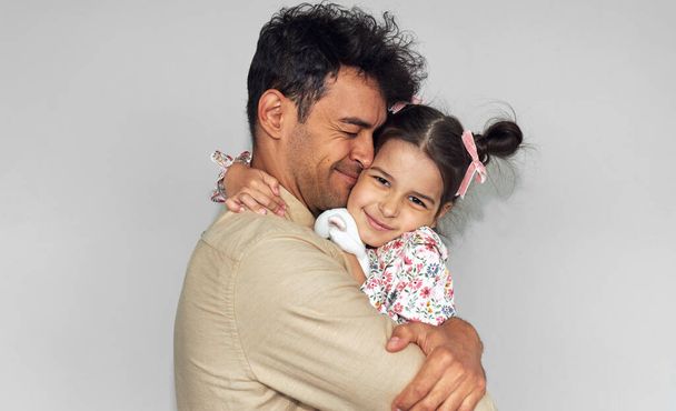 Loving father hugging his daughter against the gray wall. Happy child and daddy feeling joyful. Handsome dad and his cute little girl embraces together, shares love on Father's Day. - Foto, Imagem