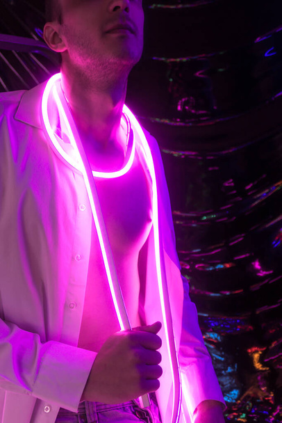 Porträt des jungen, gutaussehenden Mannes im ungeknöpften weißen Hemd. Neonfarbenes Zimmer. Der Retro-Stil der alten 90er Jahre. Reflex von Neon-Wörtern in der Nähe von Männern. - Foto, Bild