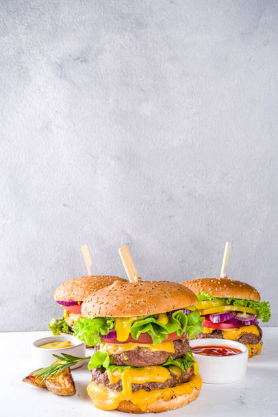 Smaczne grillowane domowe cheeseburgery z wołowiną, pomidorami, serem, pomidorami i sałatą. Na białym tle betonowym, z sosami. Góra widok miejsca kopiowania - Zdjęcie, obraz