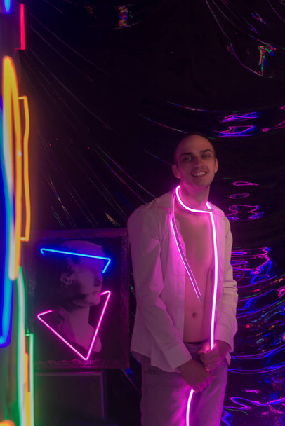 Portrait of the Young Handsome man in an unbuttoned white shirt. Neon Colored Room. Old 90's retro style. Reflect of neon words near male. - Photo, Image