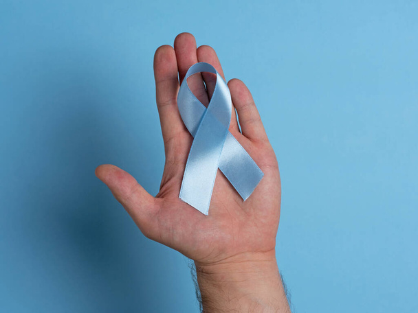 明るい青色の背景に青い前立腺がんの認識リボンを保持するクローズアップ男性の手。医療・ヘルスケアの概念 - 写真・画像