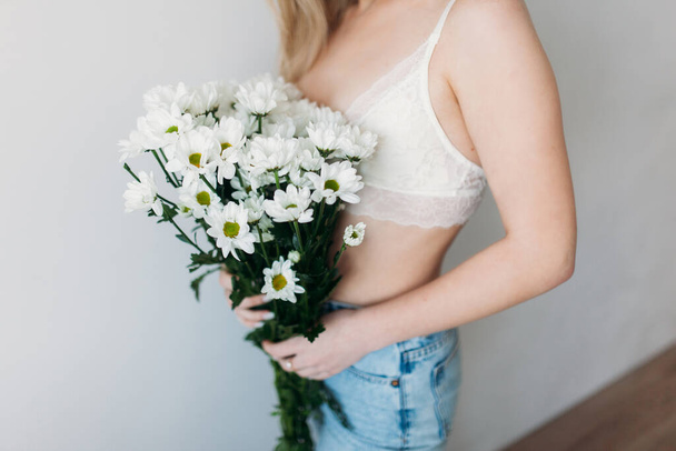A beautiful girl with a covered face with light hair in her underwear holds a bouquet of daisies. A festive bouquet of daisies in the focus of the lens. A gift for women's day or mother's Day. - Foto, imagen