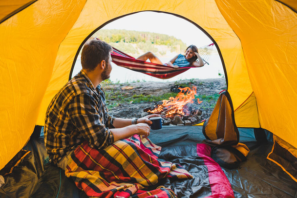 couple resting near fire. man sitting in tent woman laying on hammock - Zdjęcie, obraz