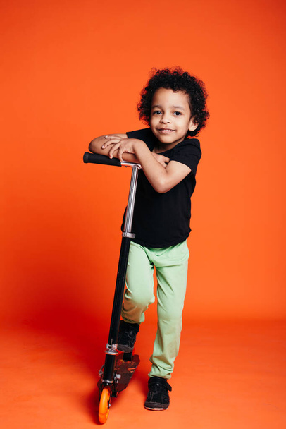 Full-length portrait of an African American boy riding a scooter in a studio on an orange background - Photo, Image