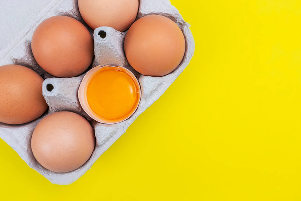 A package of six brown eggs with one opened and orange yolk on yellow background - Fotó, kép