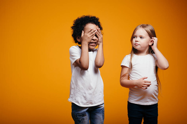Ένα αγόρι και ένα κορίτσι διαφορετικών εθνικοτήτων με λευκά μπλουζάκια παίζουν κρυφτό μαζί. Τα παιδιά δεν έχουν ρατσισμό. - Φωτογραφία, εικόνα