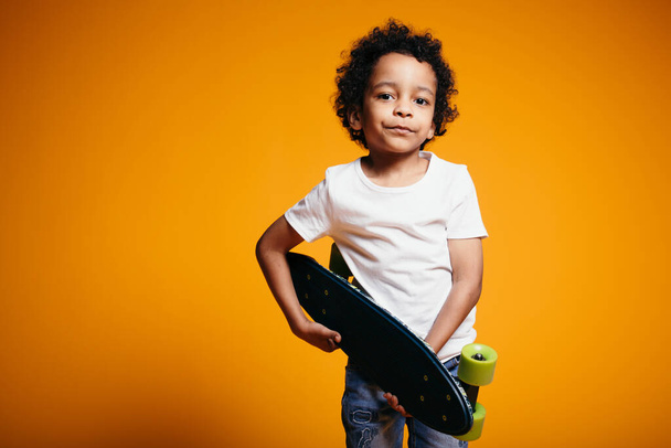 Кінкі Мараканський хлопчик у білій футболці і джинсах обіймає скейтборд і притискає його до грудей. Фотографія дітей у студії - Фото, зображення