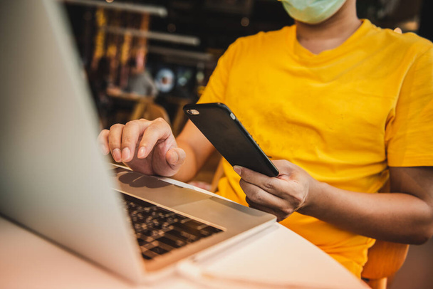 Een man in geel shirt en medisch masker met laptop en smartphone terwijl hij in coffeeshop zit - Foto, afbeelding