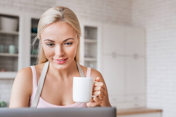 Közelkép a mosolygós nő csésze kávé és laptop a konyhában, hogy szünetelteti a főzést - Fotó, kép