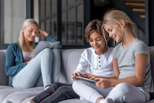 Kids using digital tablet while mother reading book on sofa in living room - Foto, Bild
