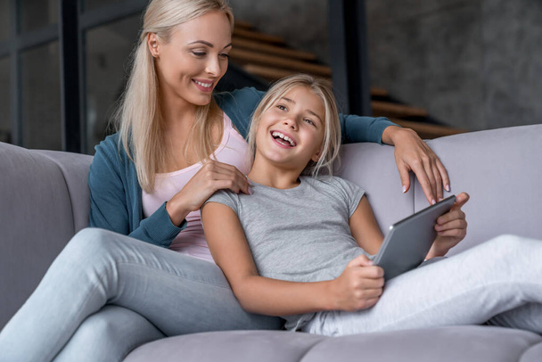 Happy mother and daughter having fun and using digital tablet sitting on sofa in living room - Foto, imagen