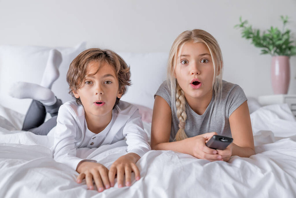 Захватывающие мальчик и девочка в пижаме лежат в спальне дома и смотрят кино.
 - Фото, изображение