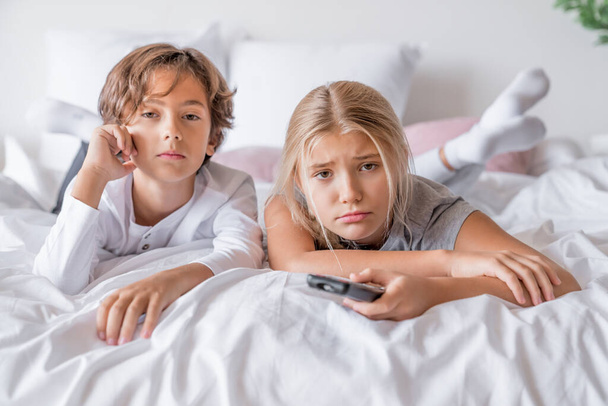 Boring boy and girl in pajamas lying in bedroom at home watching tv - Фото, изображение