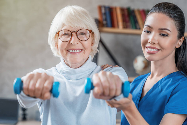 Old woman training with physiotherapist using dumbbells at home - Fotoğraf, Görsel