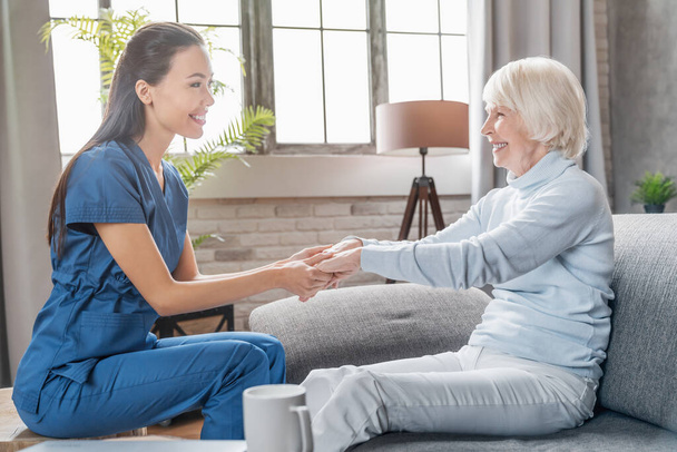 Assisting senior people. Female caregiver holding elderly woman's hands indoors - Фото, изображение