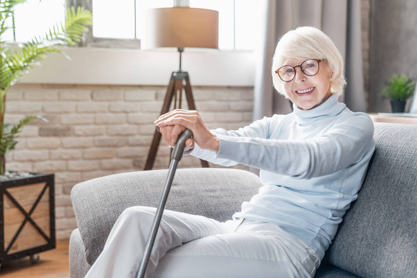 Smiling mature woman in glasses holding cane while sitting on sofa at home - Φωτογραφία, εικόνα