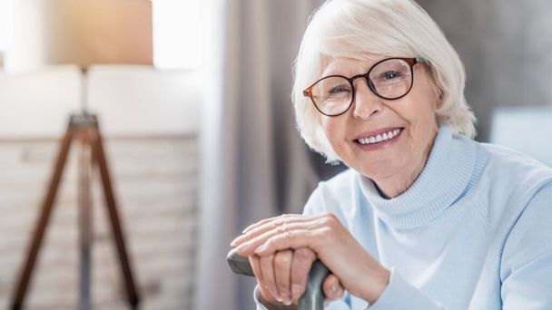 Portrait of happy mature woman in eyeglasses holding cane while sitting on sofa at home - Photo, image