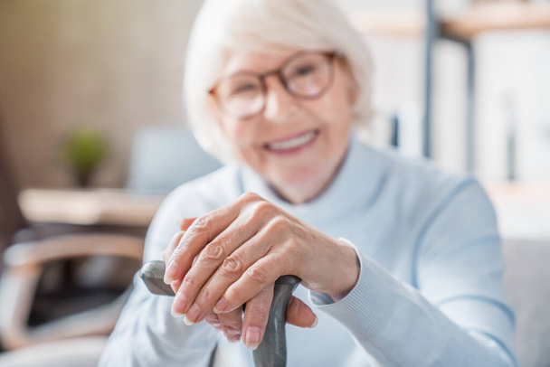 Senior woman holding cane while sitting on sofa at home with focus on hands. Old people healthcare concept - Photo, Image