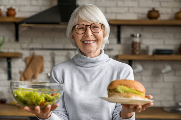 Healthy food concept. Mature smiling woman holding salad and burger - Fotografie, Obrázek