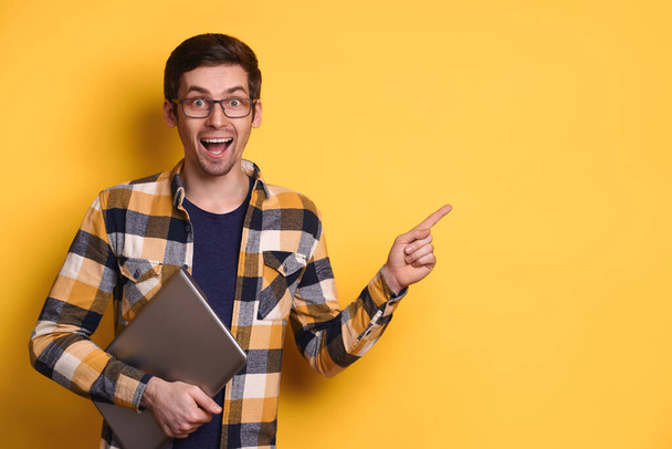 Aufgeregter junger Mann mit Brille lächelt glücklich, hält Laptop in der Hand und zeigt mit dem Zeigefinger auf freie Leerfläche für Anzeigentext. WOW, OMG, unglaublich, Promotion-Konzept - Foto, Bild