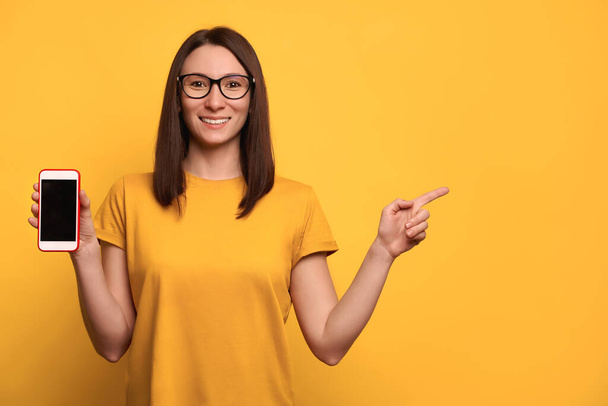 Smiling dark haired female in eyeglasses and yellow t-shirt holding cellphone with blank display, smiles and points at copy space, isolated over yellow background. Promotion concept. Sales consultant - Foto, imagen