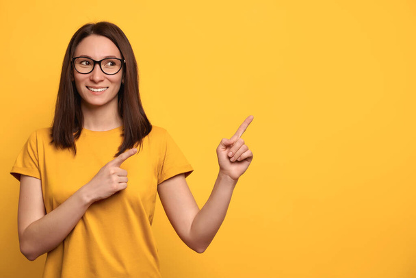 Happy young mixed race woman in glasses giving direction for advertising with index fingers, isolated on yellow background. Brunette female pointing at free space. Advert concept - Foto, Imagen