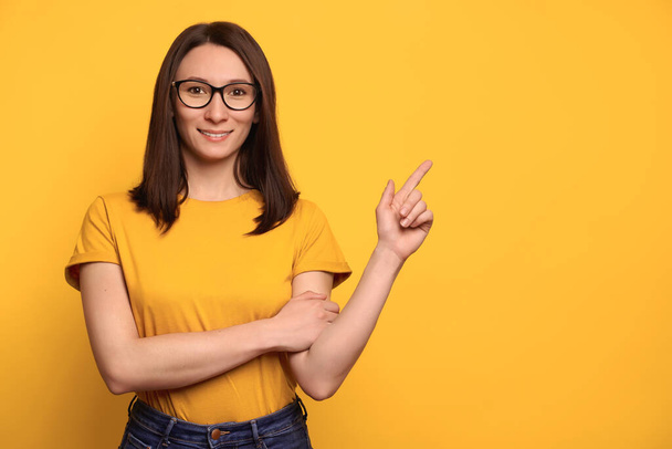 Beautiful smiling girl in eyeglasses pointing  with index finger at copy space for advertisement, isolated on yellow background. Pretty girl showing recommendation, advising product. Promotion concept - Foto, imagen