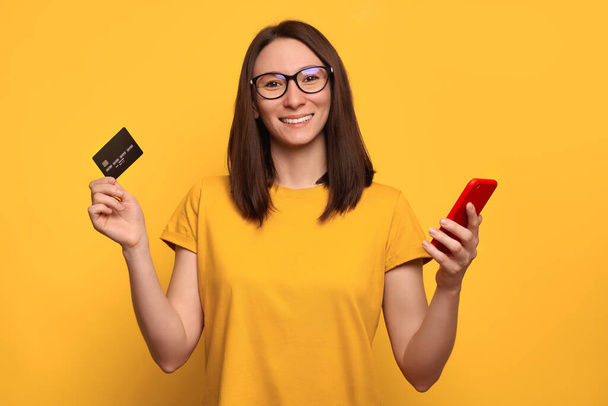 Портрет улыбающейся красивой женщины в очках с кредиткой и смартфоном на желтом фоне. Использование мобильного телефона для онлайн-банкинга, покупок и оплаты счетов. Технологическая концепция
 - Фото, изображение