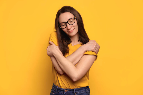 Young beautiful brunette woman in eyeglasses wearing casual yellow t-shirt hugging herself, looks calm and positive, smiling happily, love her body. Body positive, self acceptance, self esteem concept - Photo, Image