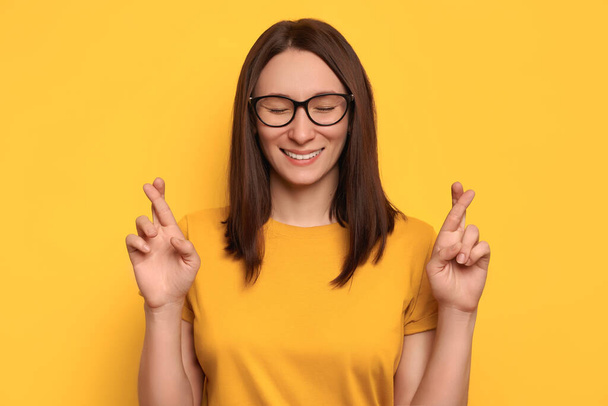 Close-up portrait of hoping beautiful woman crossing her fingers, eyes closed, asking better, smiling, wishing good luck, isolated on yellow wall background. Emotion, feeling, believe and hope concept - Fotografie, Obrázek