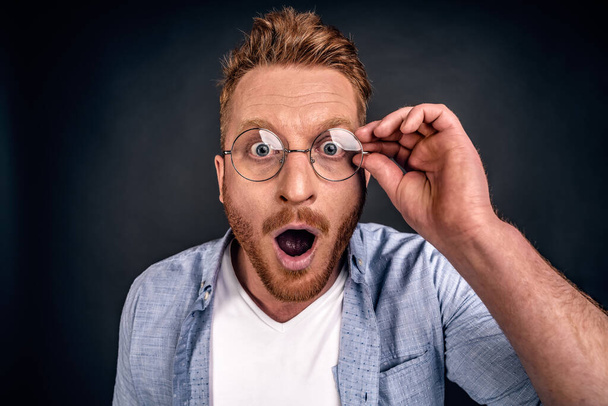 Close up portrait of stunned bearded young redhead guy drops jaw, has bugged blue eyes, sees something unbelievable and surprising, has eyewear, isolated on grey background. People, emotions concept. - Фото, зображення
