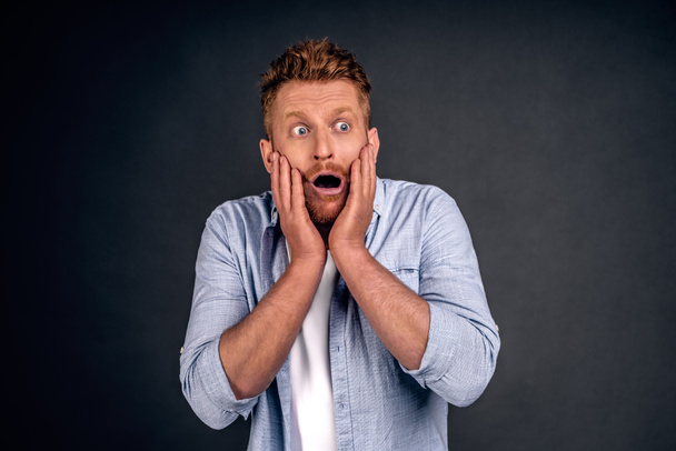 Portrait of funny handsome bearded man with red hair, expressing shock or being terrified of something, standing with opened mouth and popped eyes over gray background. Guy is pale as if he saw ghost. - Фото, изображение