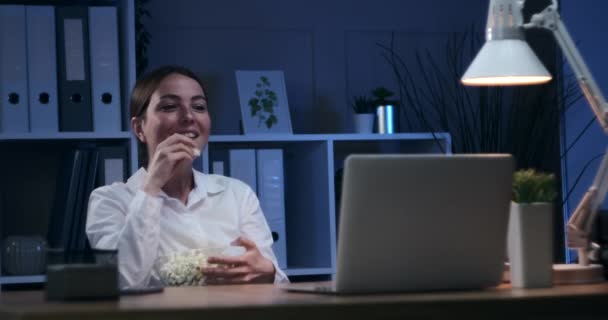 Businesswoman eating popcorn and watching movie at night office - Imágenes, Vídeo