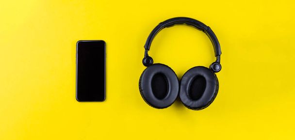 Zwarte koptelefoon en mobiele telefoon op een gele achtergrond. Concept van muzikale draadloze streaming, luisteren naar muziek door alles heen. - Foto, afbeelding