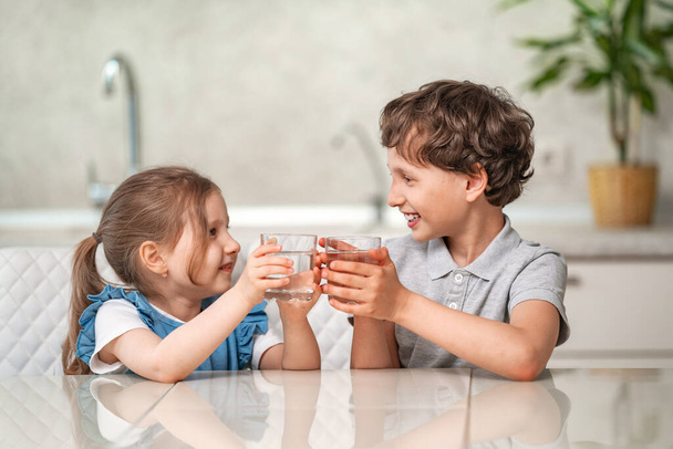 Funny little children drink water in the kitchen at home. The boy and girl are smiling happily, holding a glass of clear water and looking at each other. Water balance. Prevention of dehydration - 写真・画像