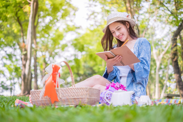 Усміхаючись азіатською жінкою, яка відпочиває на пікніку обід на відкритому повітрі, вона сидить на траві і читає книжку. - Фото, зображення