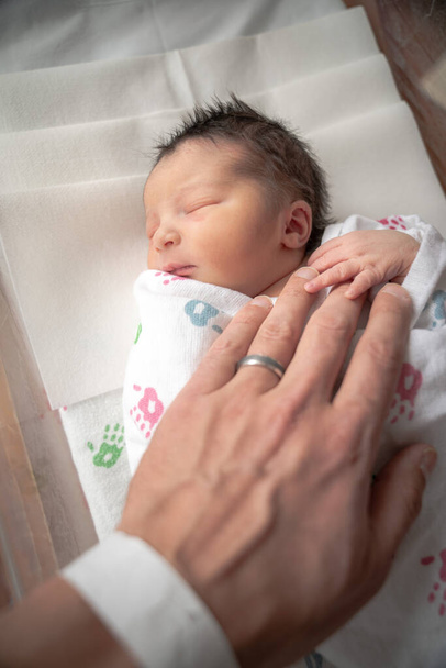A newborn infant baby girl in a blanket swaddle rests her tiny hand and fingers on her fathers wrinkled hand as she sleeps peacefully. - Photo, Image