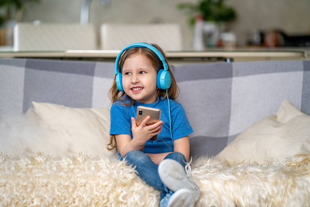 Happy little girl listening to music in blue headphones, at home. The child is sitting with smartphone in his hands and enjoys listening to his favorite songs and singing. Funny emotions. - Photo, Image