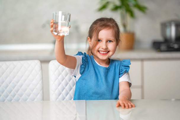 Cute little girl drinking water in the kitchen at home. The little girl is smiling happily, holding a glass of clean water and wants to drink it. Water balance. Prevention of dehydration - Foto, Imagen