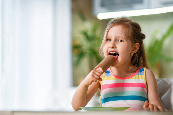 happy child is sitting at the kitchen table with ice cream on a stick in chocolate glaze raspberry cherry and strawberry filling pink. girl licking a Popsicle. sweet cool dessert on a hot day. - Photo, Image