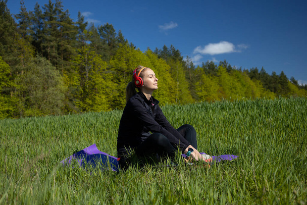 A girl sits on a fitness mat and listens to music after a workout in the open air outside the city. Green field and forest - Photo, Image