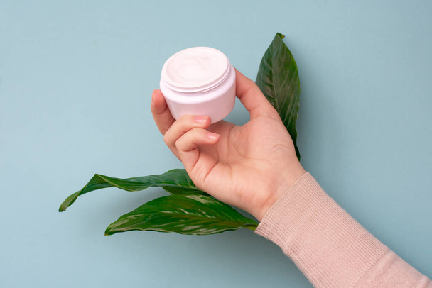 Young girls hand is holding a jar with white soft cream on pale blue background with large green leaves.Concept of eco cosmetic and beauty procedure. - Foto, immagini