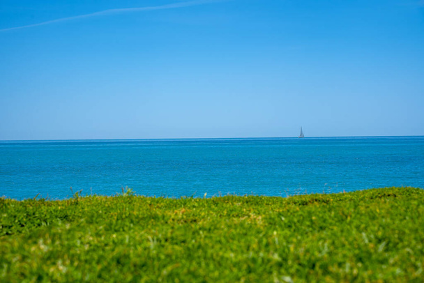 Blauwe zeegezicht met zeilboot aan de horizon. Uitzicht vanaf de groene heuvel van een eiland. - Foto, afbeelding