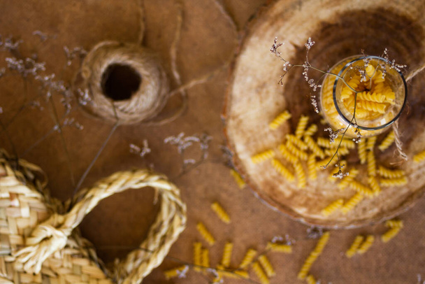 Pastas espirales en un frasco de vidrio sobre un soporte de madera, junto a él están las ramas secas de los árboles, sobre un fondo beige
 - Foto, Imagen