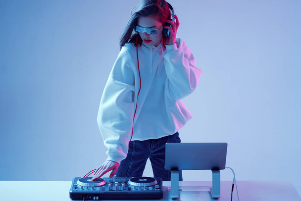 Cool young girl DJ mixes music on a mixing console and laptop, in stylish clothes, glasses on a neon background. - Fotó, kép