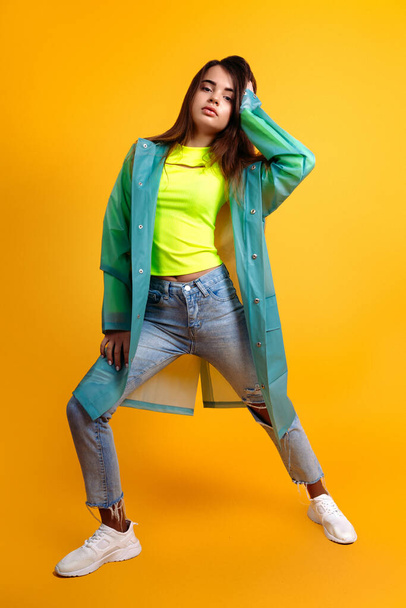 Young teenager girl dressed in jeans and a raincoat in the studio on a yellow background - Foto, afbeelding